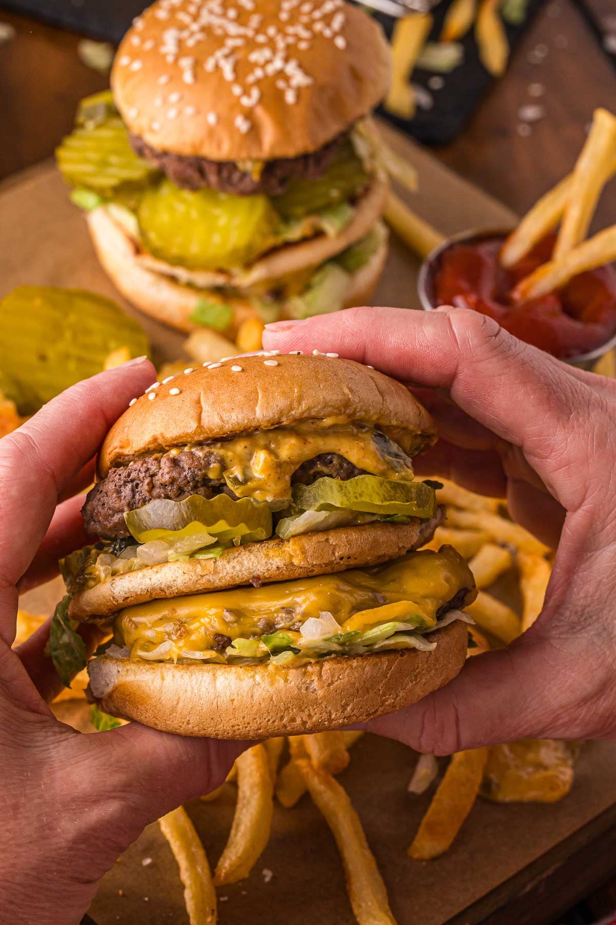 Two hands hold a double cheeseburger with pickles, lettuce, and melted cheese. Another similar burger, French fries, and a container of ketchup are on the table in the background.