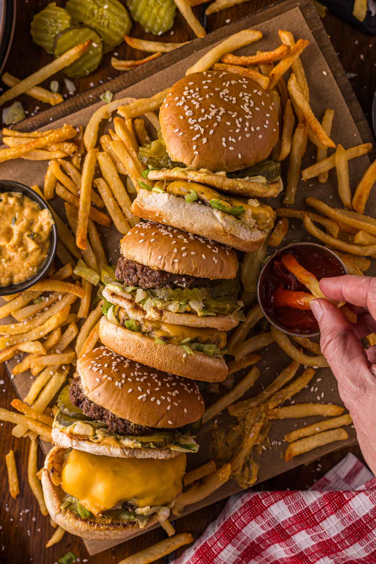Four cheeseburgers with lettuce and pickles are arranged in a row on a board surrounded by French fries, with a hand dipping fries into ketchup.