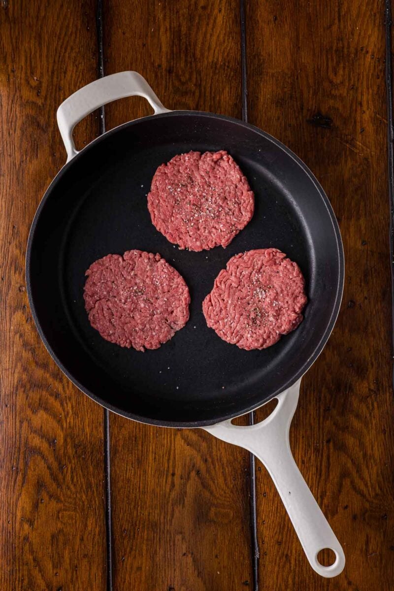 Three raw hamburger patties seasoned with pepper are arranged in a black skillet on a wooden surface.