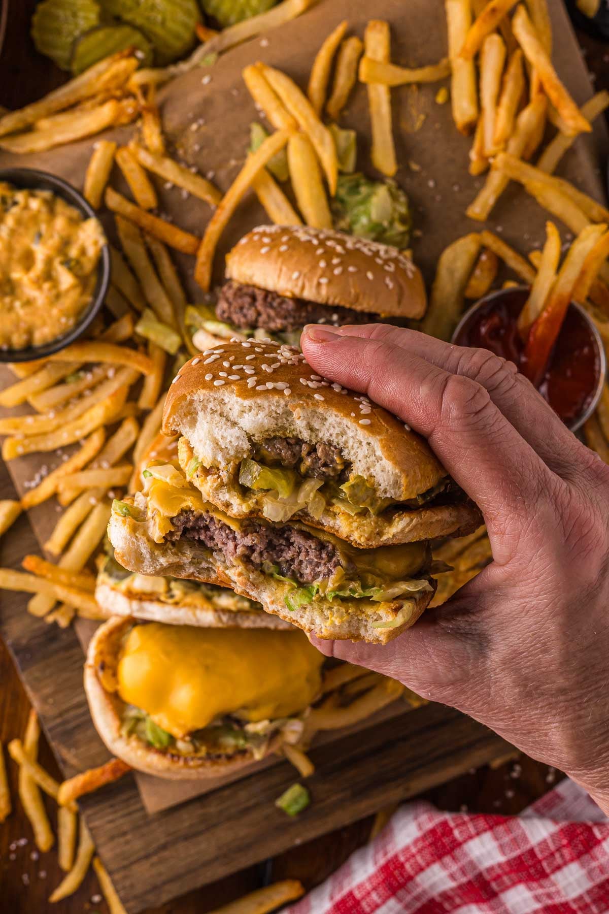 A hand holding a half-eaten cheeseburger over a table with French fries, more burgers, pickles, cheese dip, and ketchup.