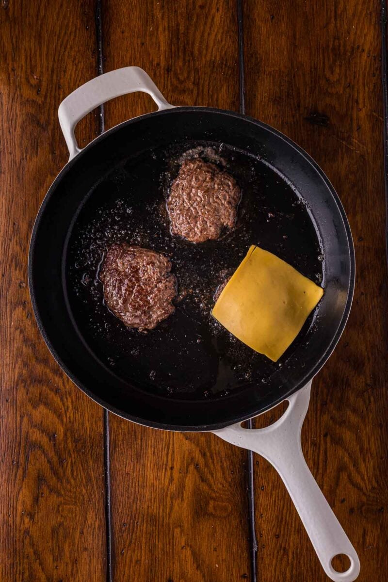A white skillet on a wooden surface contains three burger patties, one topped with a slice of cheese.