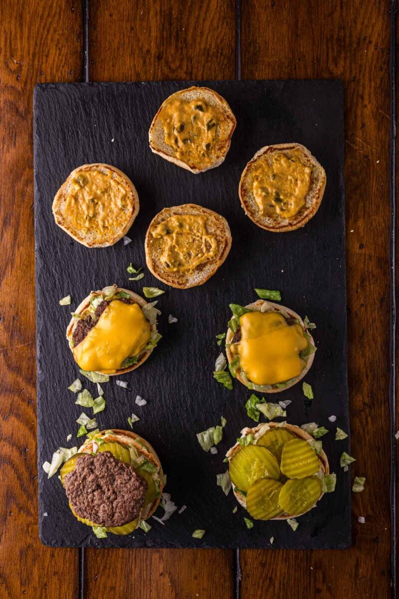 Overhead view of three open-faced cheeseburgers on a black slate board, each topped with different ingredients; the top bun halves are spread with sauce.
