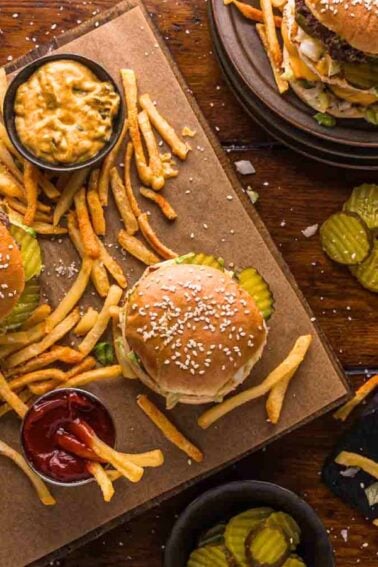 Three sesame seed burgers with lettuce and cheese served on boards with French fries, pickles, cheese dip, and ketchup on a wooden table.