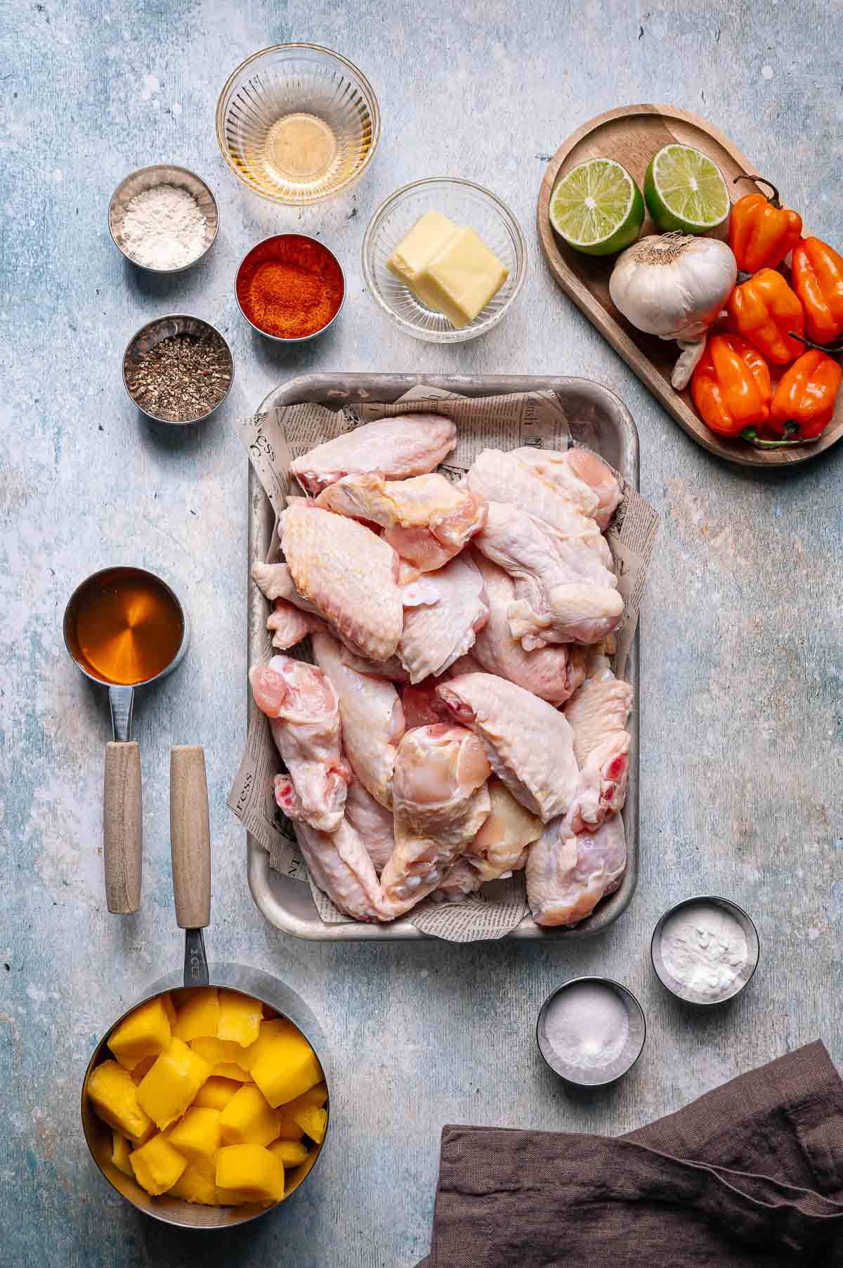 Raw chicken wings on a tray surrounded by small bowls of spices, butter, lime, garlic, habanero peppers, honey, flour, and diced mango on a light surface.