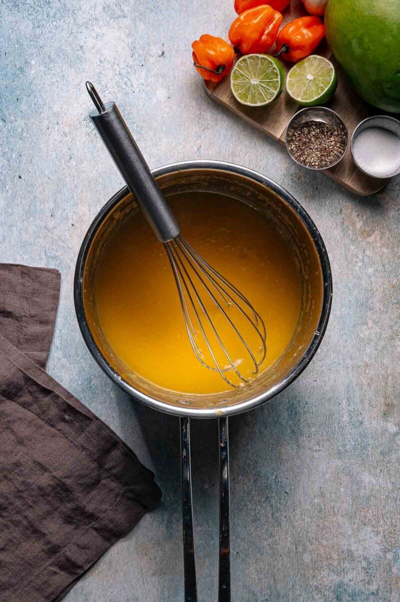 A saucepan filled with yellow sauce and a whisk, next to a brown cloth, with limes, habanero peppers, salt, and pepper on a tray nearby.