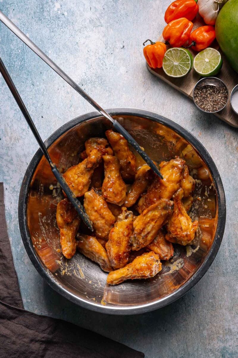 Tongs mixing glazed chicken wings in a metal bowl, with limes, black pepper, garlic, and habanero peppers on a tray nearby.