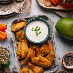 A tray of glazed chicken wings garnished with green onions is served with a bowl of ranch dip; fresh vegetables, spices, and lime slices surround the tray.