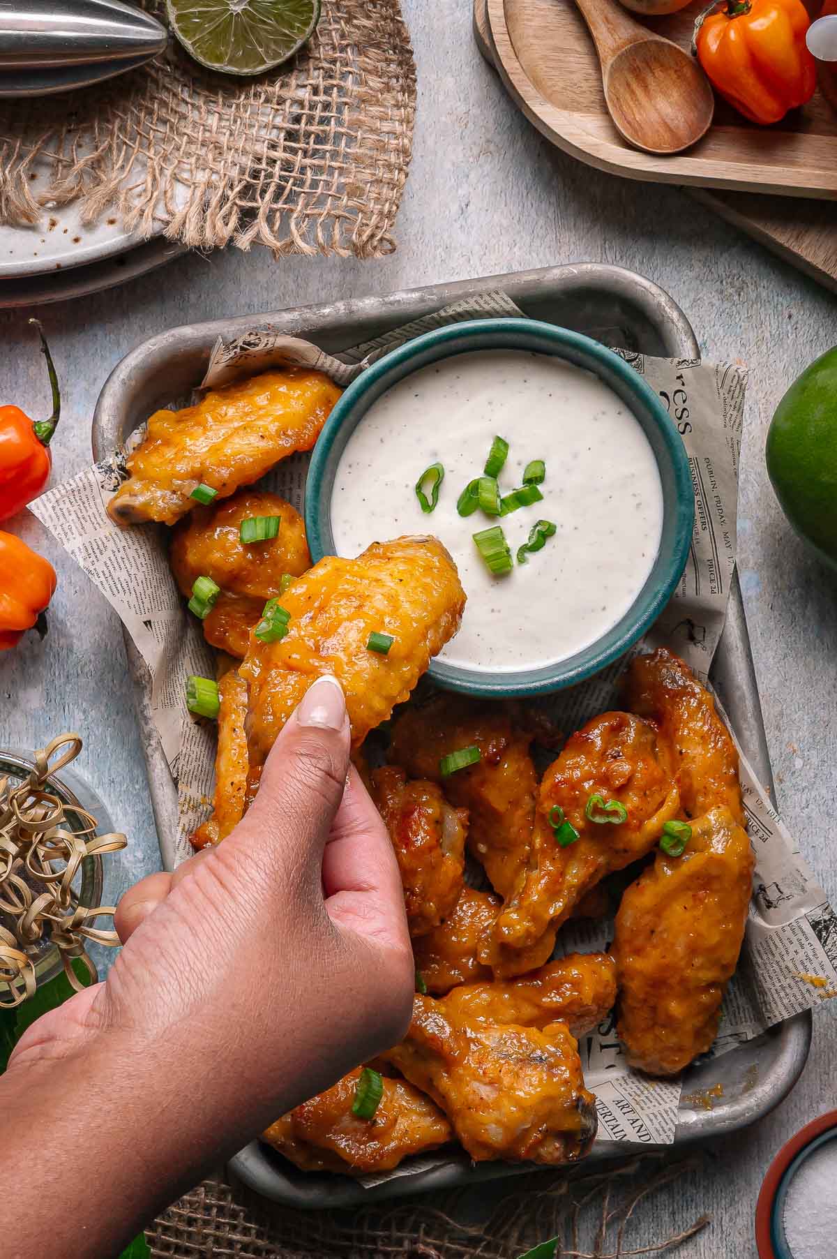 A hand dips a sauced chicken wing into a bowl of ranch dressing. More wings, garnished with chopped green onions, are arranged on a tray nearby.