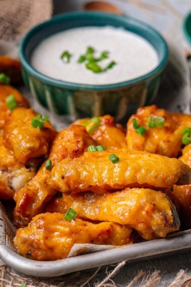 A tray of glazed chicken wings garnished with chopped green onions is served alongside a bowl of creamy dipping sauce topped with green onions.