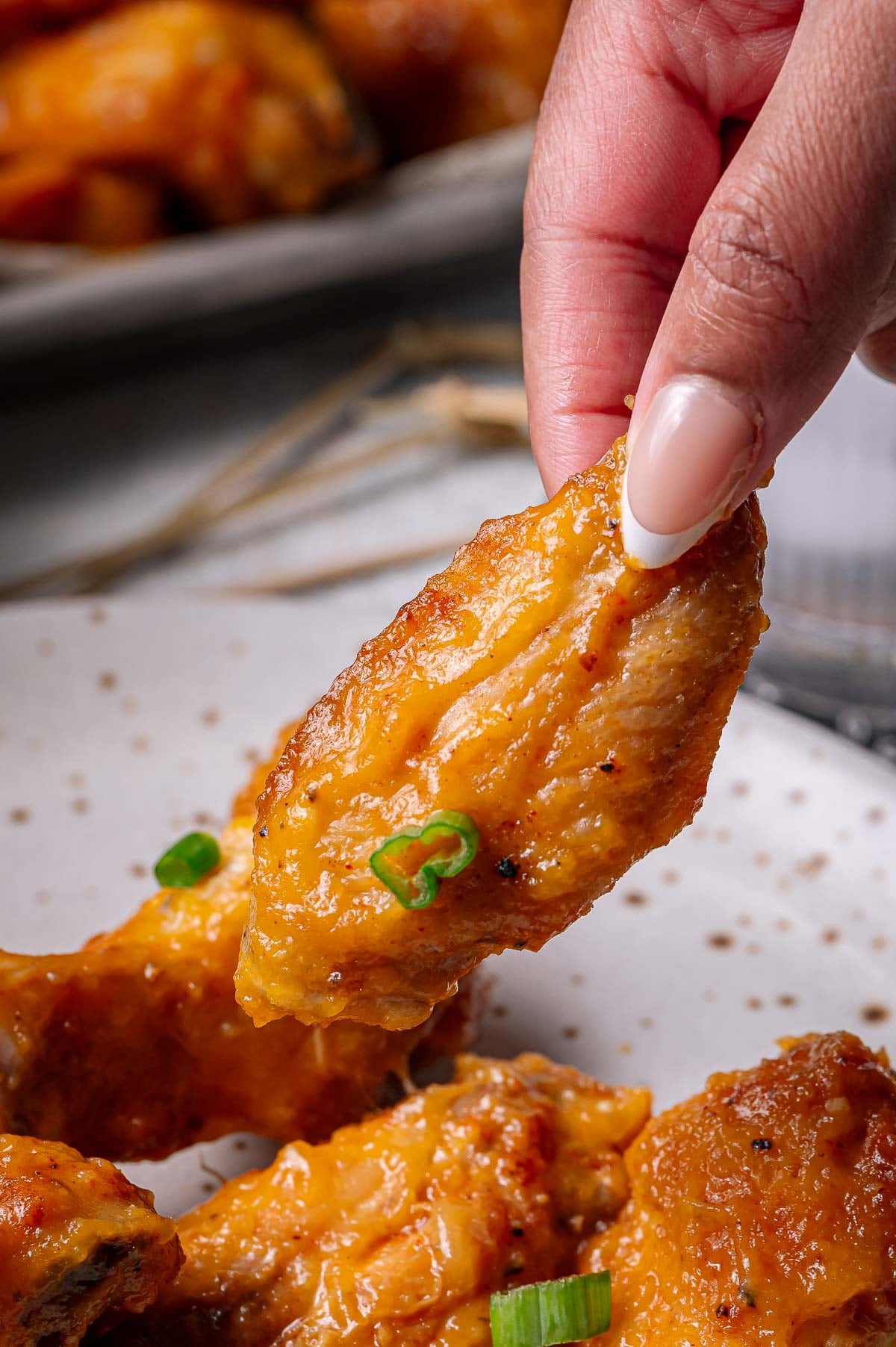 A hand holding a saucy chicken wing garnished with sliced green onion over a plate with more chicken wings.