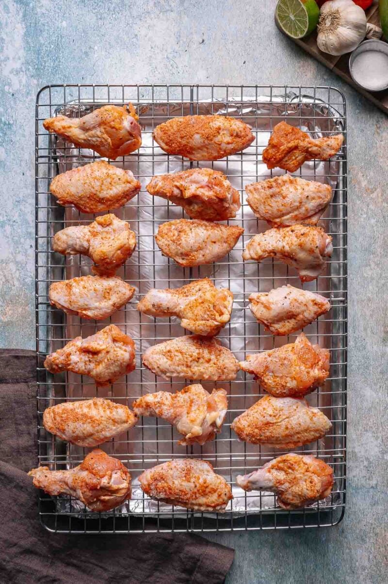 A wire rack holds 20 raw, seasoned chicken wings spaced evenly apart, ready for cooking. A dark cloth, garlic, lime, and small bowls are visible nearby.