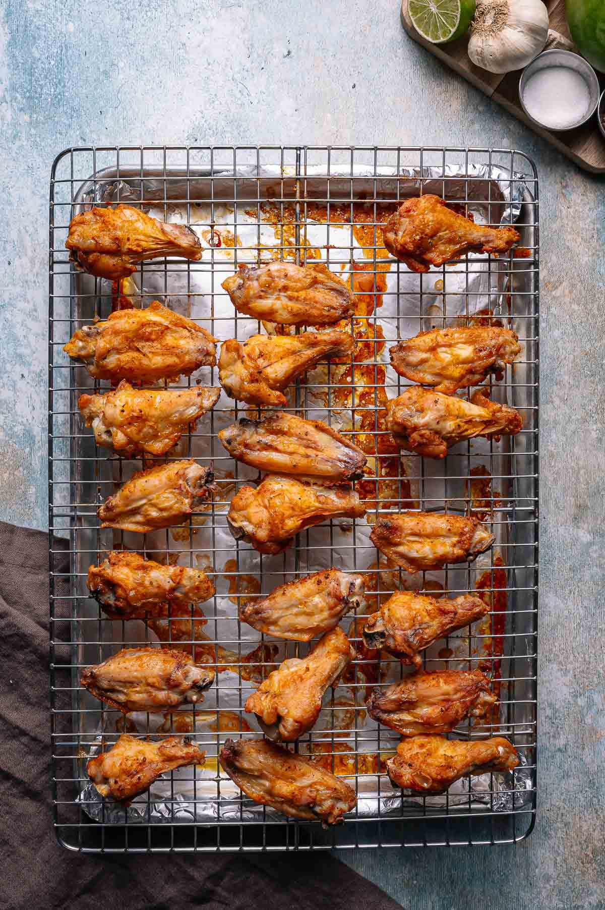A baking tray with a wire rack holds cooked chicken wings spaced evenly, with some sauce residue underneath. Garlic, lime, and salt are visible in the upper right corner.