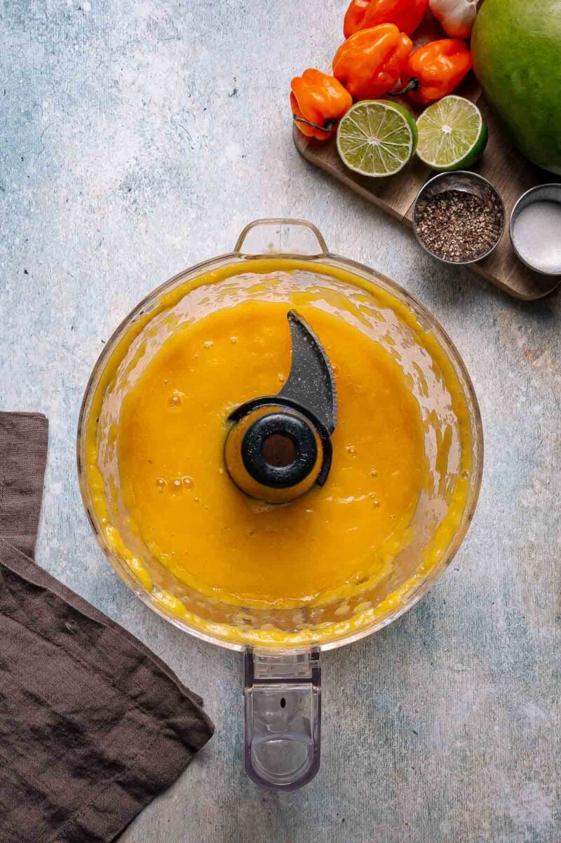 A food processor filled with blended yellow sauce sits on a countertop next to a brown towel, with limes, habanero peppers, salt, and pepper on a wooden board.