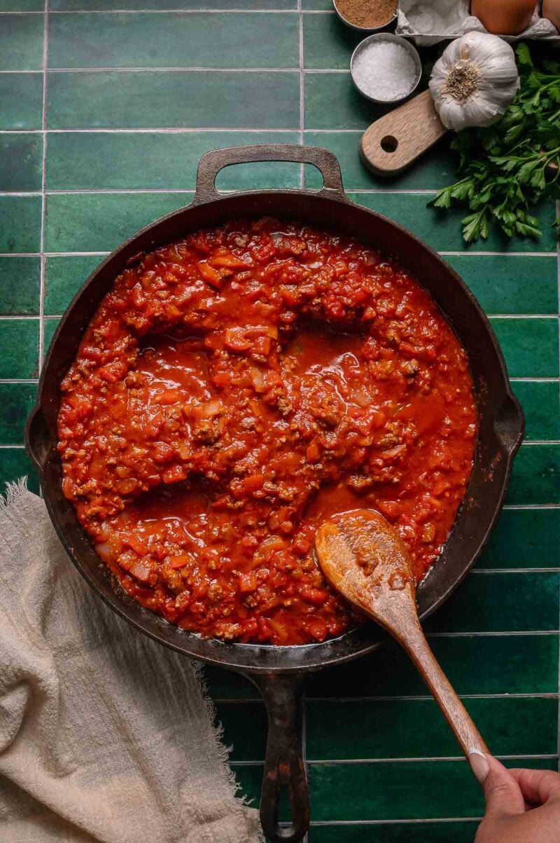 A cast iron skillet filled with chunky shakshouka sauce sits on a green tiled surface, with a wooden spoon and fresh ingredients nearby.
