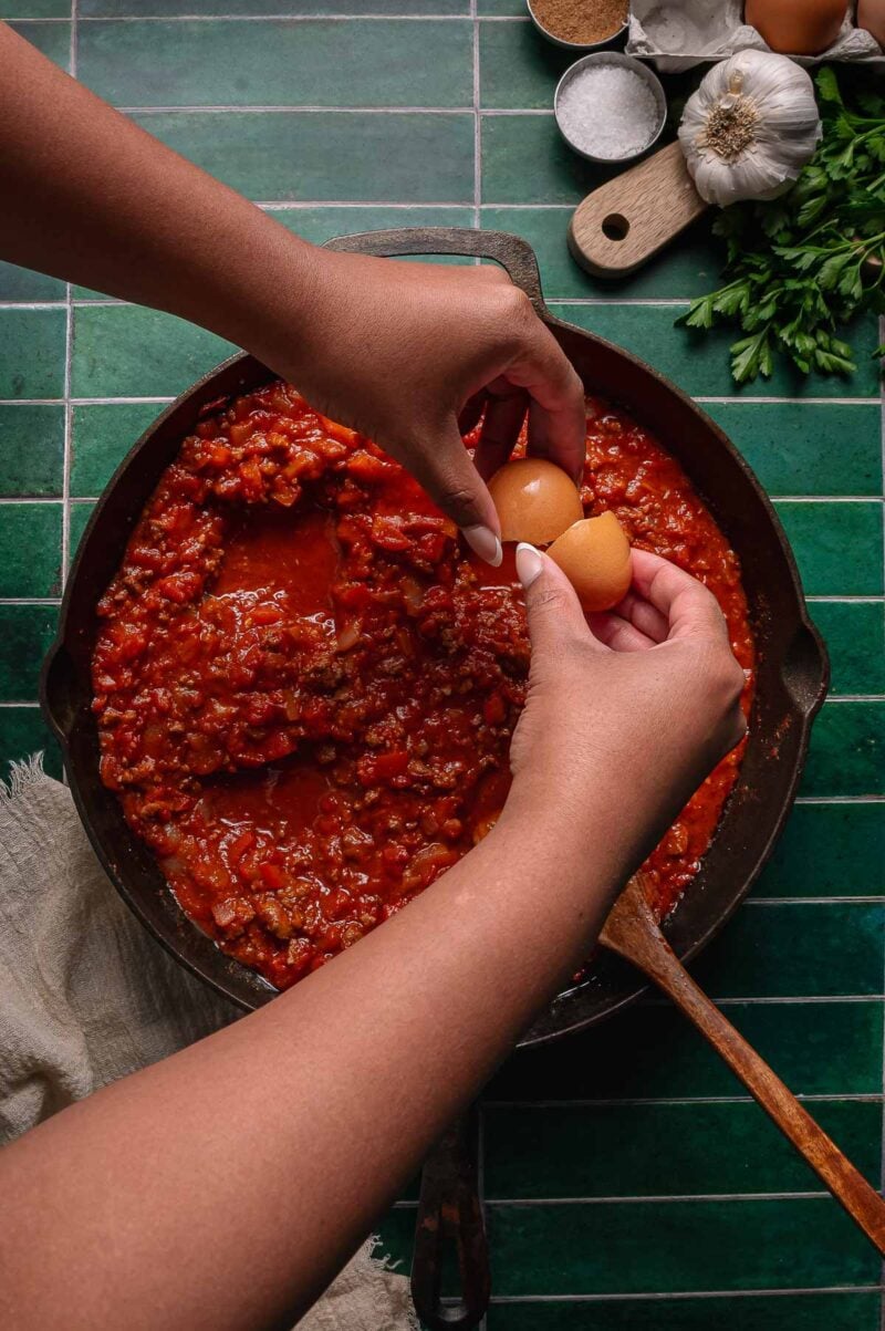 A person cracks an egg into a cast iron skillet filled with shakshuka sauce and ground lamb, with various ingredients on the side.
