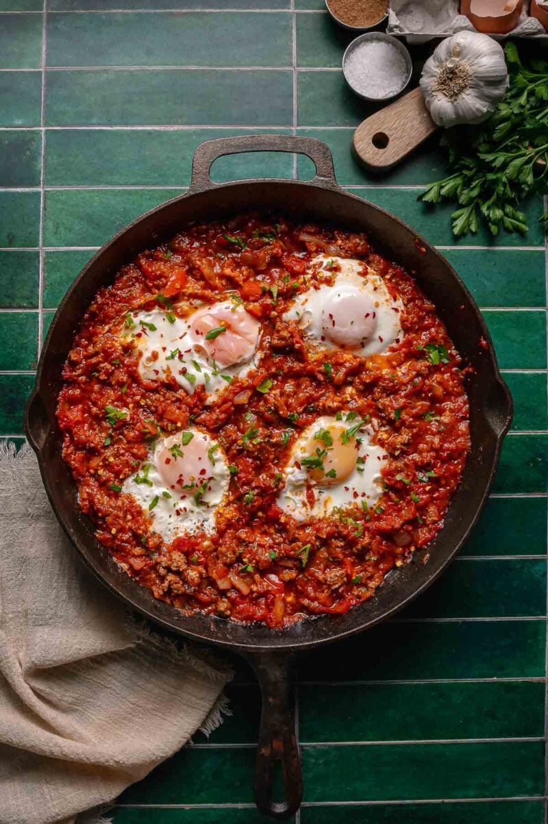 A cast iron skillet filled with shakshuka; poached eggs in a spiced tomato sauce, garnished with herbs, on a green tiled surface with ingredients nearby.