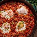 A cast iron skillet filled with poached eggs in a chunky red shakshuka sauce, garnished with chopped herbs and red pepper flakes.