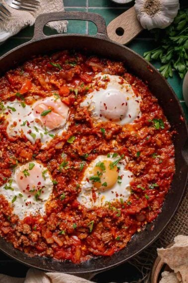 A cast iron skillet filled with poached eggs in a chunky red shakshuka sauce, garnished with chopped herbs and red pepper flakes.
