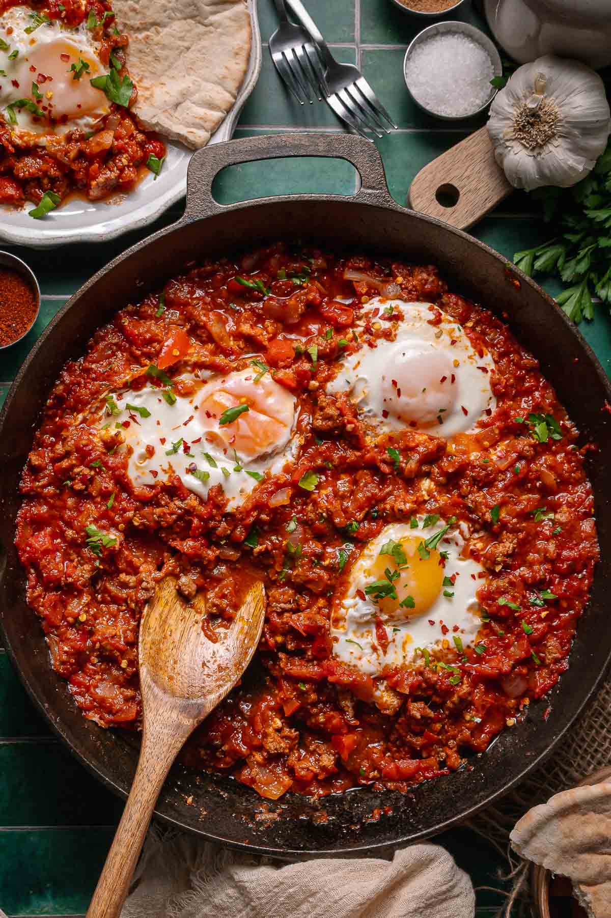 A skillet of shakshuka with poached eggs, garnished with herbs, sits on a table with pita bread, garlic, and seasonings nearby.