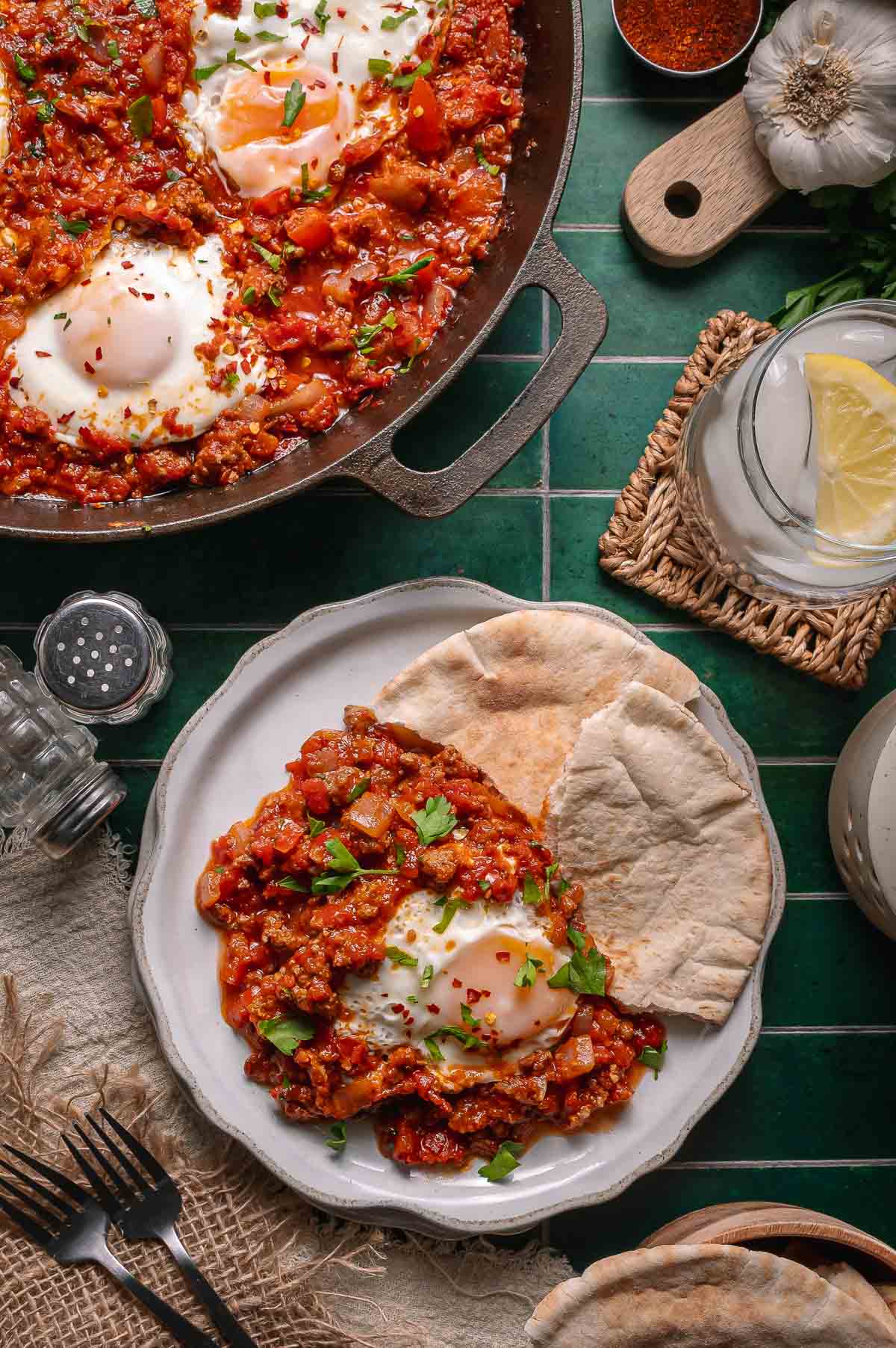 A plate of shakshuka with two pieces of pita bread, next to a pan of shakshuka, a glass of water with lemon, and utensils on a green-tiled surface.