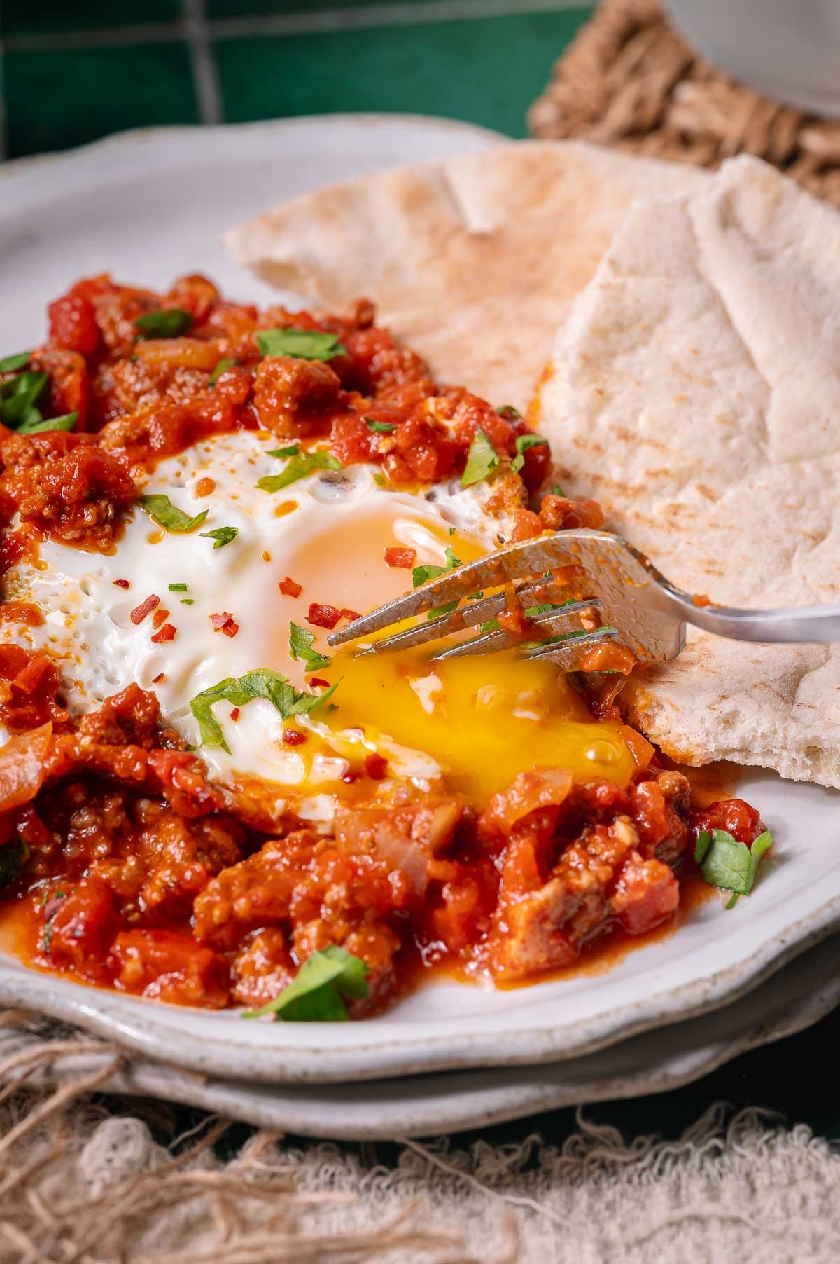 A plate with poached eggs in a tomato-based sauce, garnished with herbs, served with pita bread. A fork is cutting into the egg, releasing the yolk.
