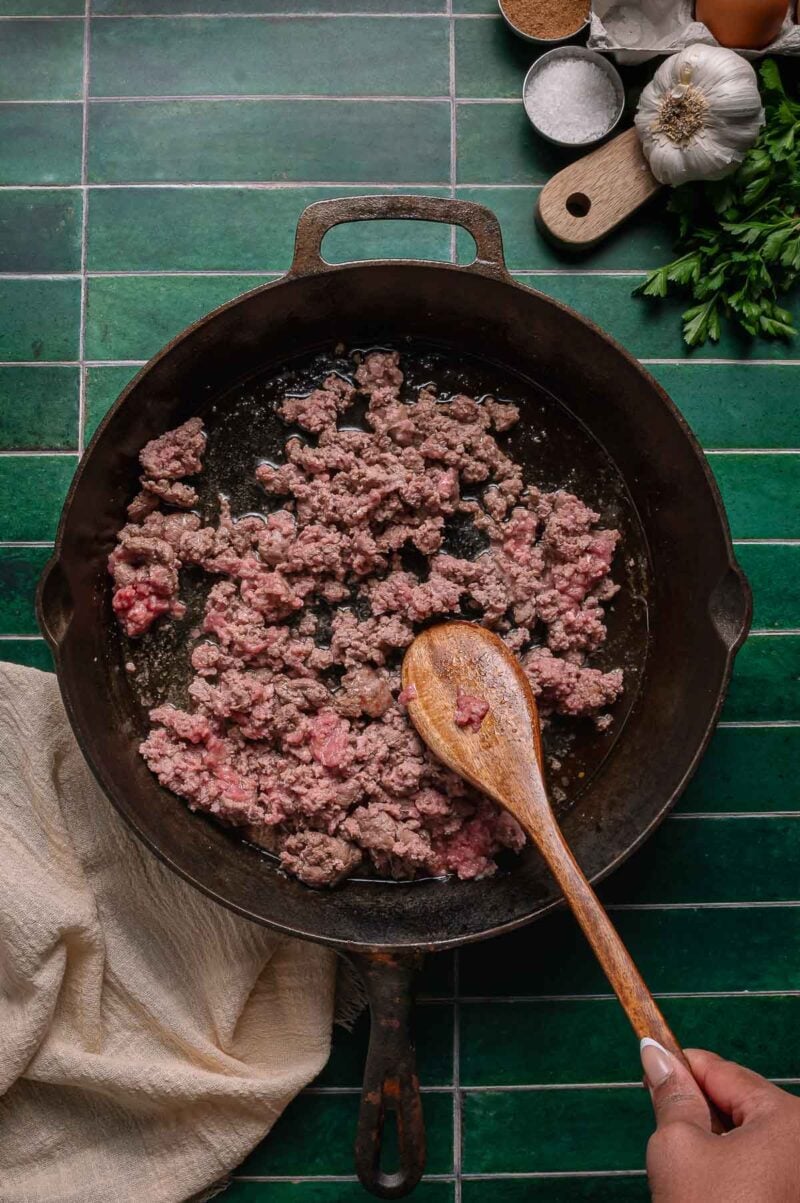 Ground lamb is being cooked in a cast iron skillet and stirred with a wooden spoon; seasonings, garlic, and parsley are visible in the background.