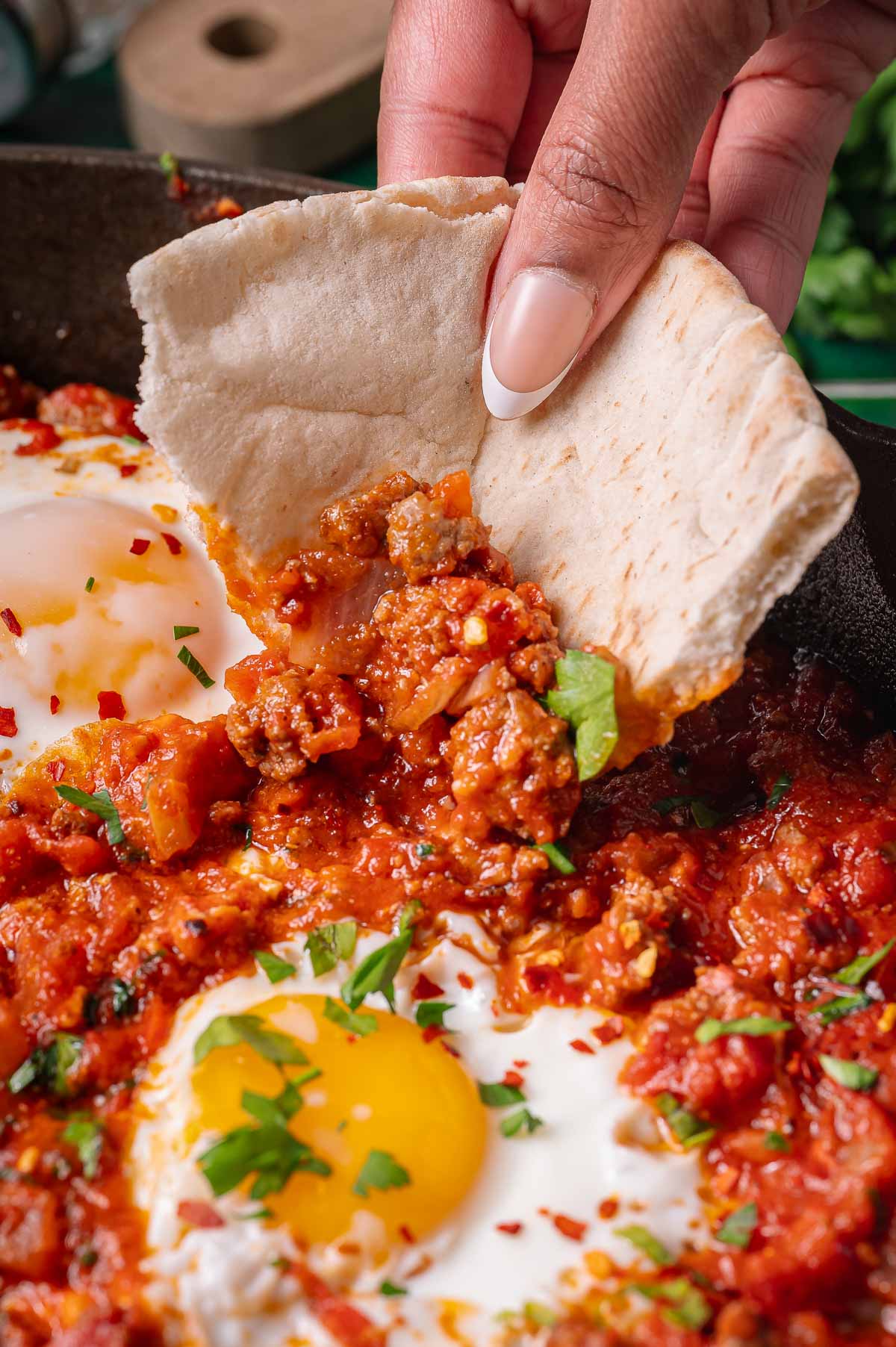 A hand scoops tomato-based shakshuka with ground meat and a poached egg using a piece of pita bread.