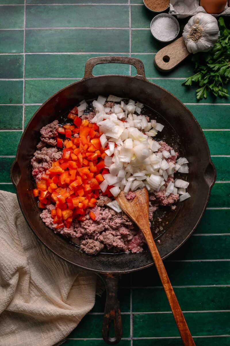 A cast iron skillet on a green tiled surface contains ground lamb, diced red bell pepper, and chopped onion being sautรฉed with a wooden spoon.