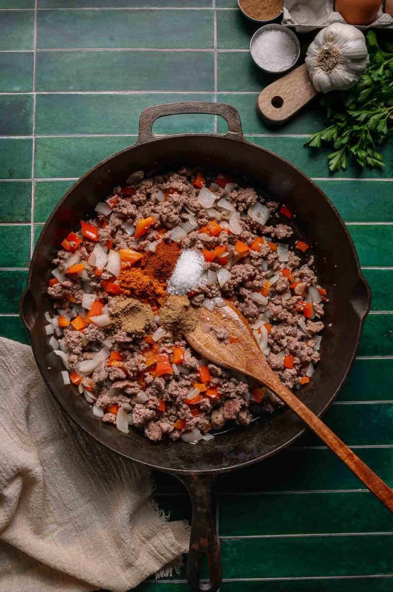 A cast iron skillet with ground lamb, chopped onions, red bell pepper, and spices being mixed with a wooden spoon on a green tiled surface.