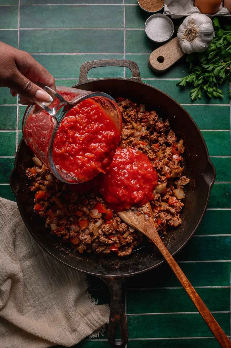 A hand pours chopped tomatoes from a measuring cup into a cast iron skillet with cooked ground lamb and vegetables on a green tiled countertop.