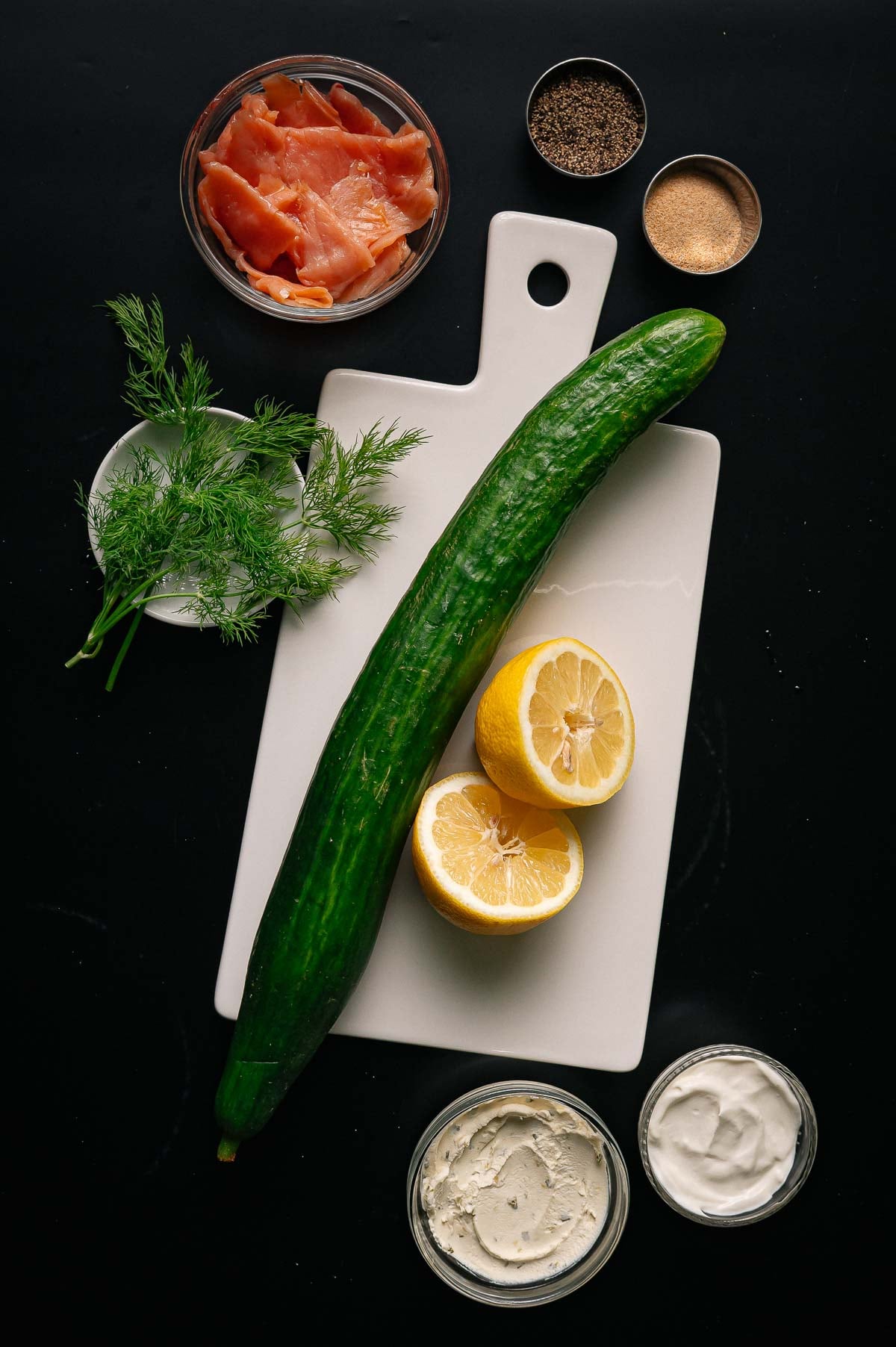 A white cutting board with a cucumber, halved lemon, fresh dill, and surrounding bowls of smoked salmon, pepper, garlic powder, cream cheese, and sour cream on a black surface.