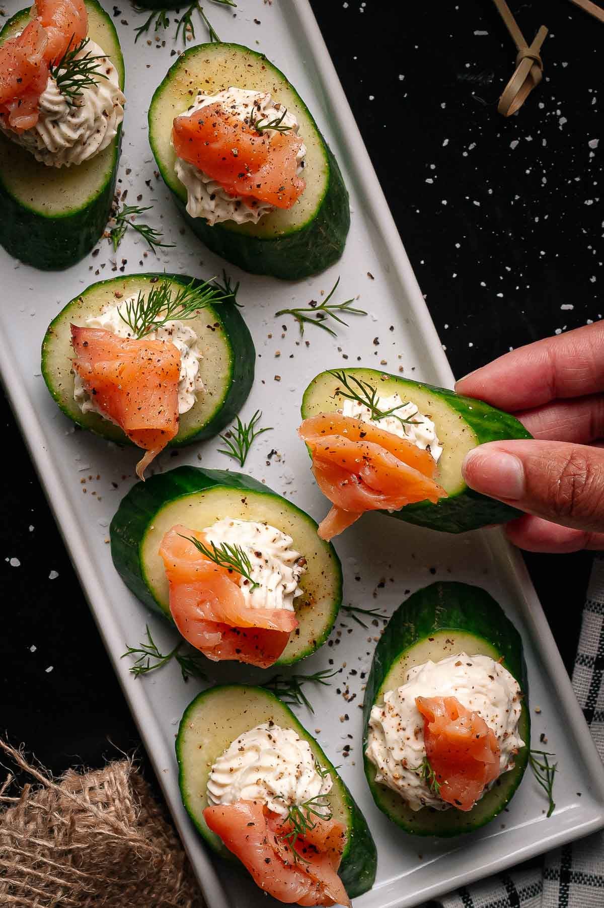 A hand picking up a cucumber slice topped with cream cheese, smoked salmon, dill, and pepper from a rectangular white plate with more identical appetizers.