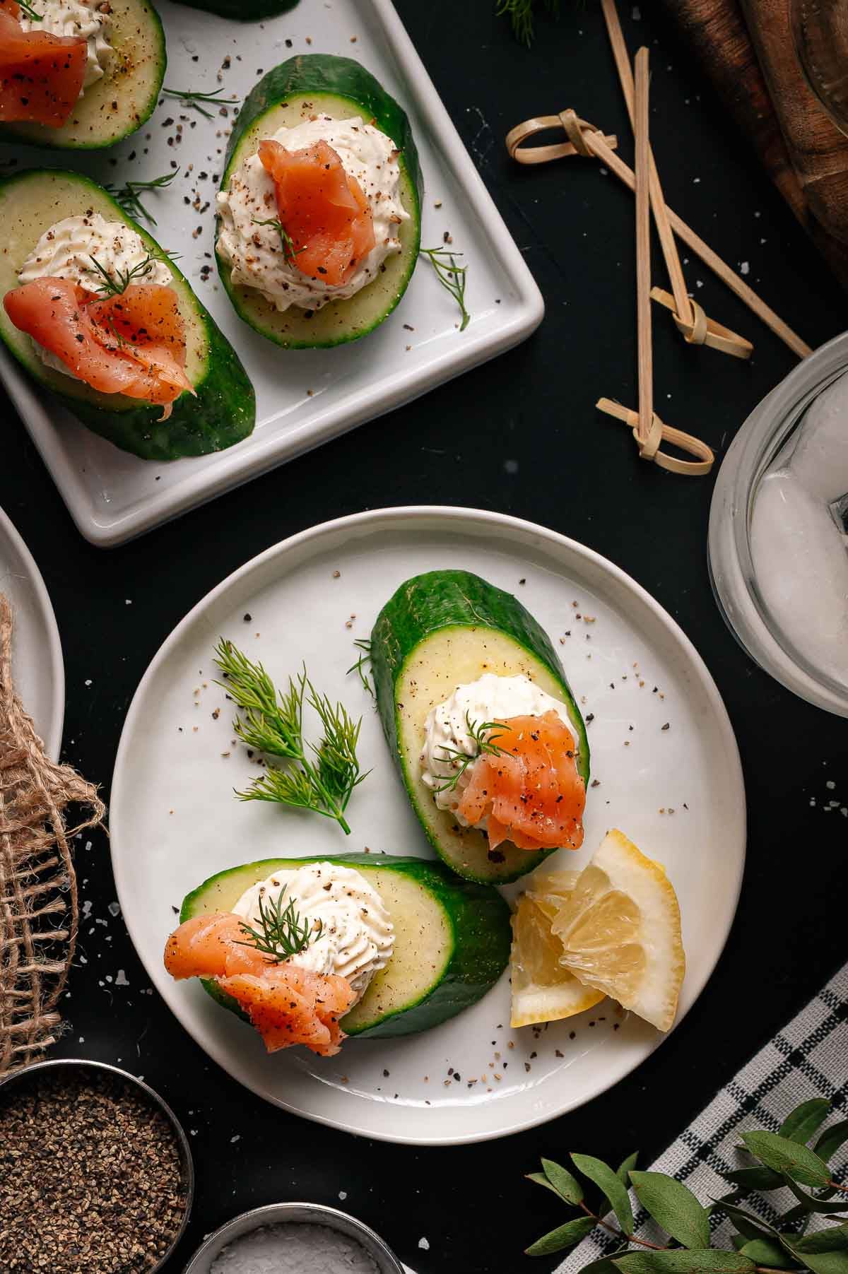 Three cucumber slices topped with cream cheese, smoked salmon, and dill are arranged on a white plate with a lemon wedge. Additional appetizers and ingredients are visible around the plate.