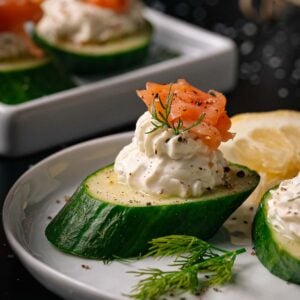 Sliced cucumber topped with a swirl of cream cheese, a piece of smoked salmon, dill, and cracked pepper, served on a white plate with a lemon wedge.