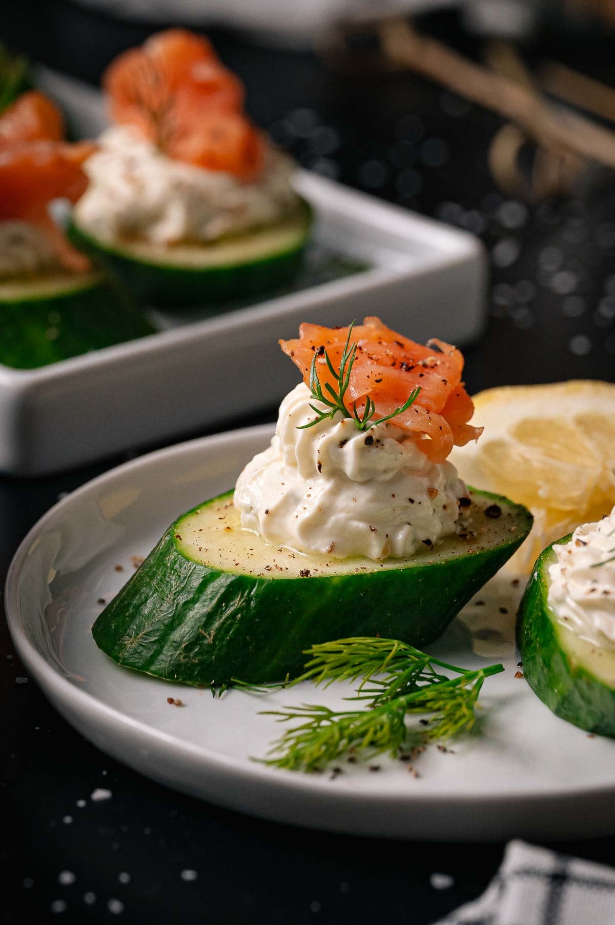 Sliced cucumber topped with a swirl of cream cheese, a piece of smoked salmon, dill, and cracked pepper, served on a white plate with a lemon wedge.