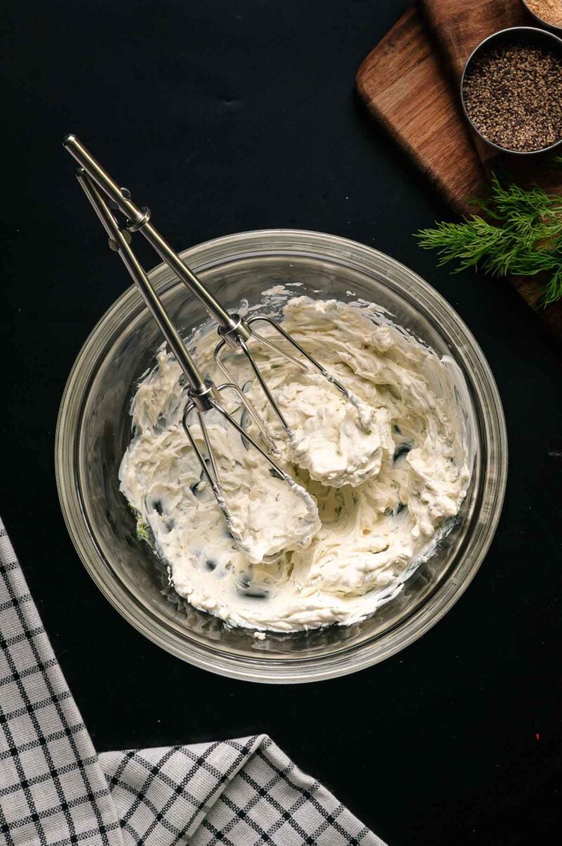 A glass bowl of whipped cream cheese mixture with herbs sits on a dark surface beside a hand mixer, a checked towel, a cutting board, dill, and a metal dish of ground pepper.