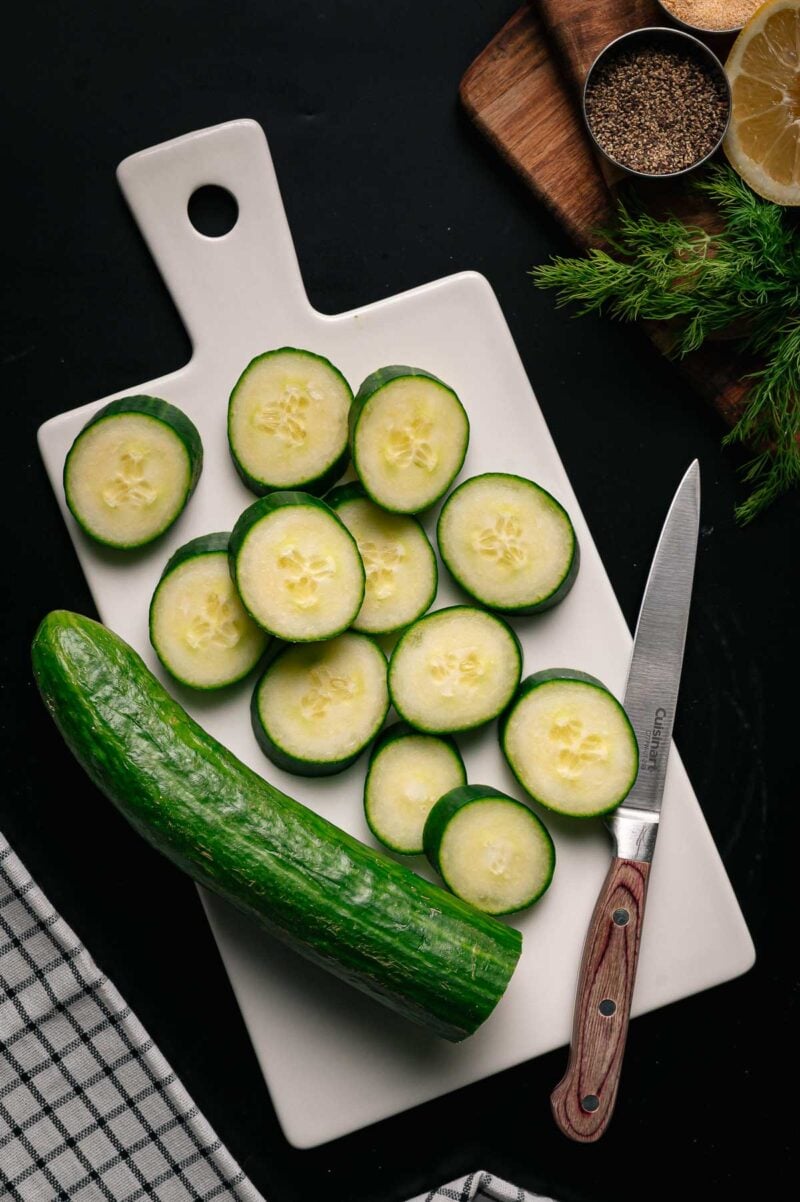 Sliced cucumber arranged on a white cutting board with a knife, whole cucumber, and herbs on a dark surface.