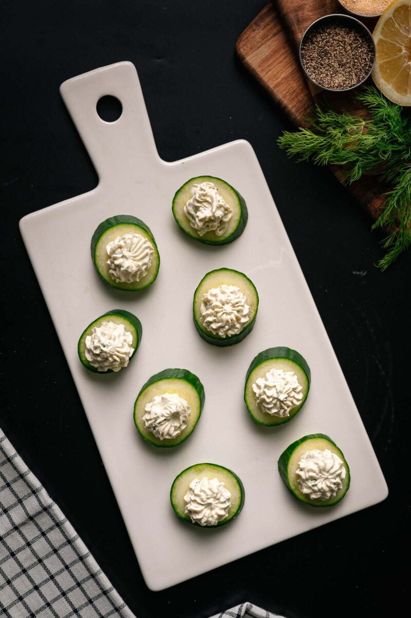 Eight cucumber slices topped with piped cream cheese are arranged on a rectangular white cutting board, with herbs, pepper, and lemon visible in the background.