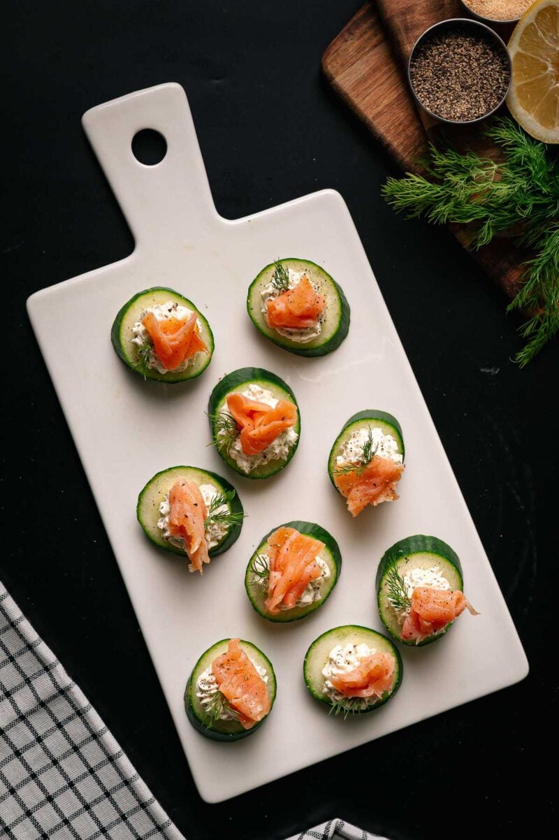 Nine cucumber slices topped with cream cheese, smoked salmon, and dill are arranged on a white rectangular serving board. A towel, lemon, dill, and black pepper are nearby.