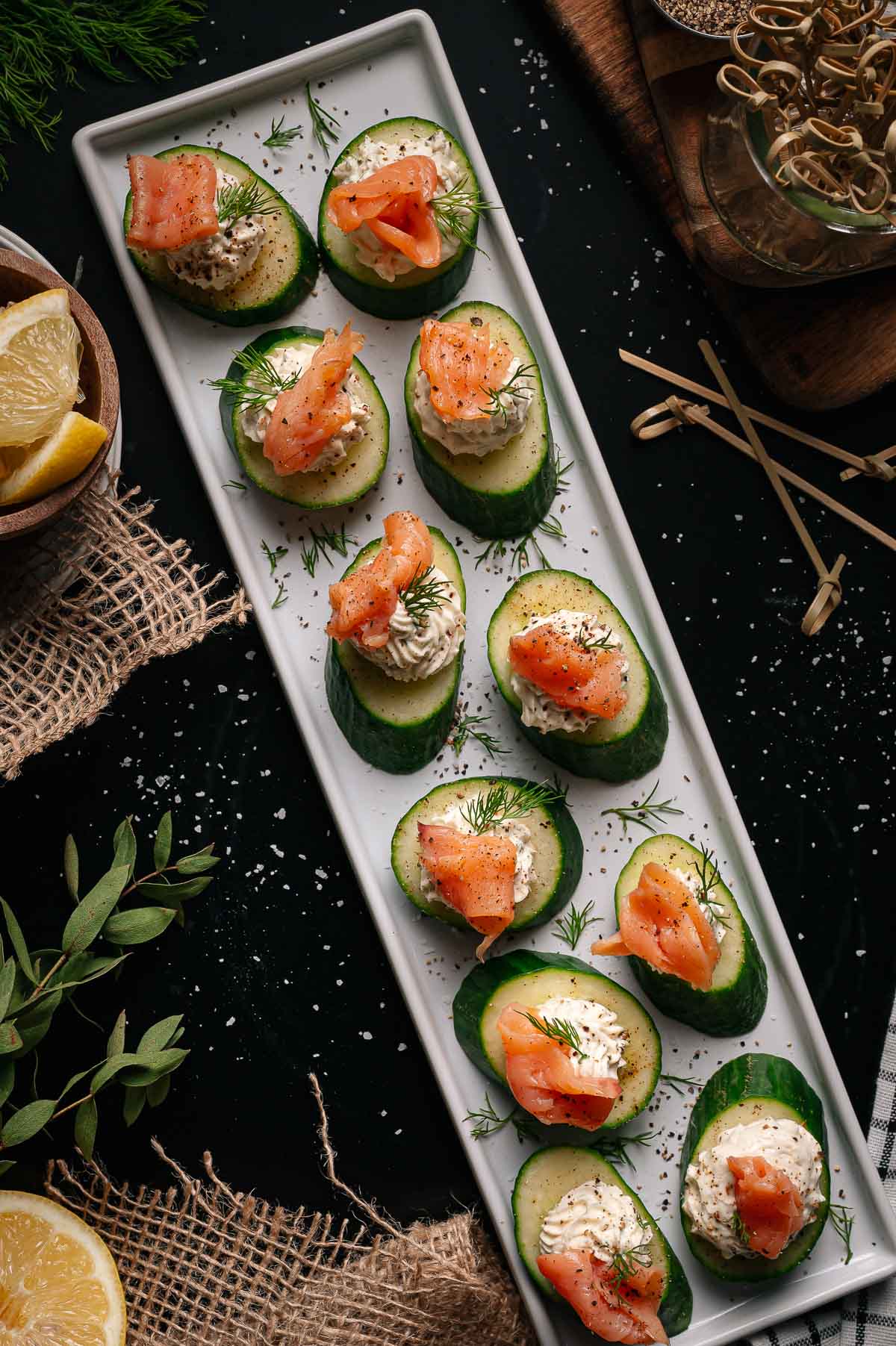 Sliced cucumbers topped with cream cheese, smoked salmon, and dill are arranged on a rectangular white platter. Lemons, herbs, and wooden picks are nearby.