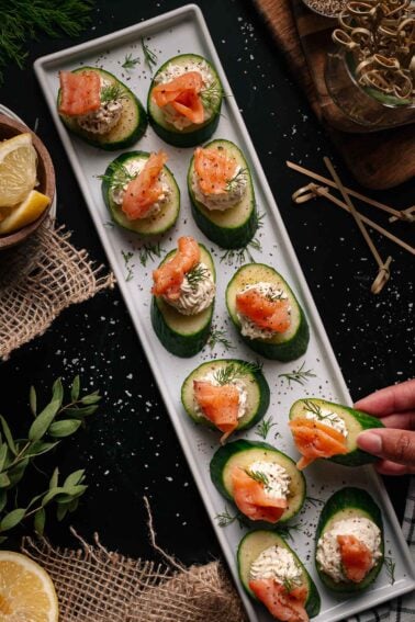 Cucumber slices topped with a creamy spread, fresh dill, and pieces of smoked salmon are arranged on a rectangular white platter. A hand is picking up one of the appetizers.