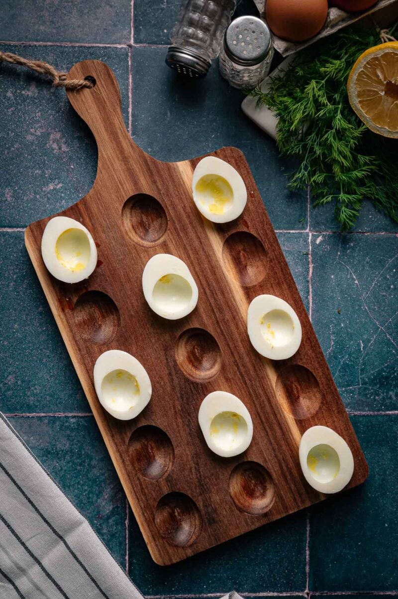 Hard-boiled egg halves are arranged on a wooden deviled egg tray, with kitchen ingredients and utensils visible in the background.