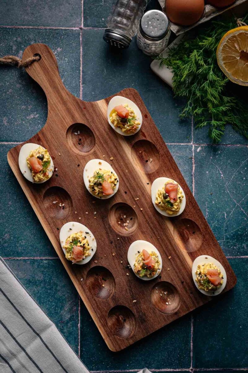 Deviled eggs topped with herbs and smoked salmon are arranged on a wooden serving board, placed on a dark tiled surface with ingredients nearby.