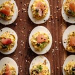 Halved hard-boiled eggs filled with a creamy smoked salmon filling, garnished with herbs, capers, seasoning, and smoked salmon, arranged on a wooden surface.