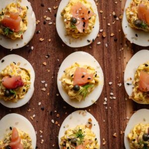 Halved hard-boiled eggs filled with a creamy smoked salmon filling, garnished with herbs, capers, seasoning, and smoked salmon, arranged on a wooden surface.