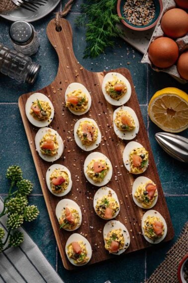 A wooden board with deviled eggs topped with smoked salmon, herbs, and seeds, surrounded by a lemon half, eggs, seasoning, and fresh dill on a dark countertop.