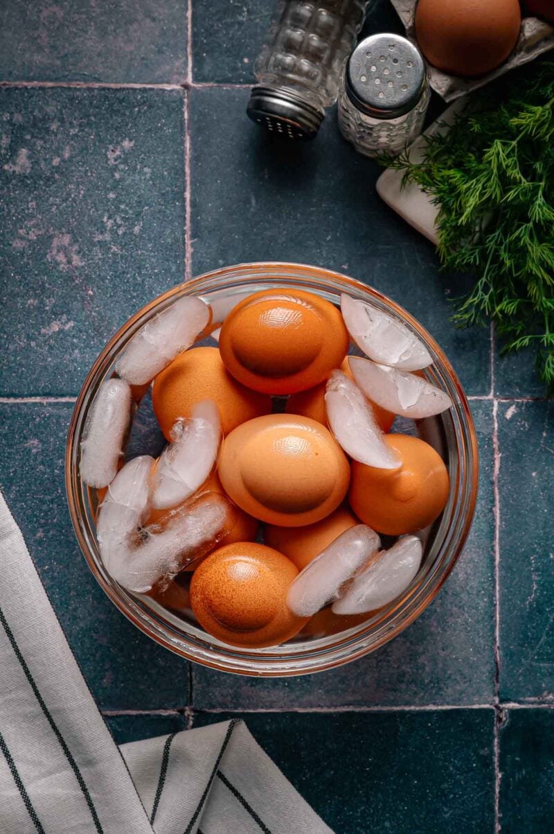 A glass bowl with brown eggs and ice cubes, set on a blue tile countertop with a striped towel, salt and pepper shakers, and fresh dill nearby.