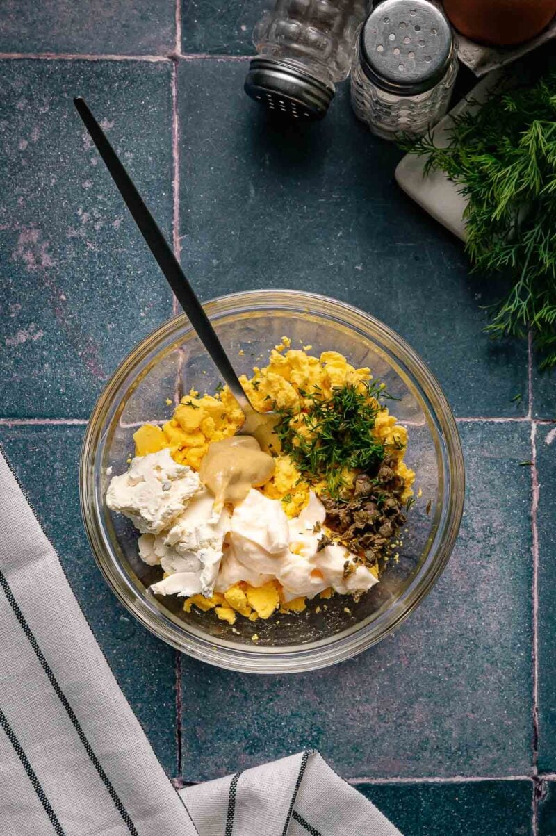 A glass bowl with chopped hard-boiled eggs, mayonnaise, mustard, fresh dill, capers, and cream cheese, with a black spoon, on a tiled surface next to salt, pepper, and fresh dill.