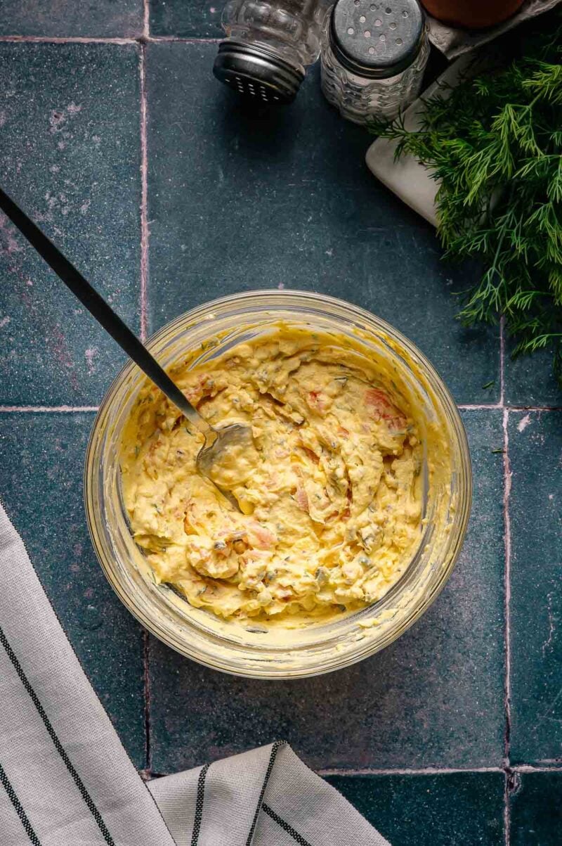 A glass bowl of deviled egg filling with a spoon on a dark tiled countertop, surrounded by fresh dill, salt and pepper shakers, and a striped cloth.