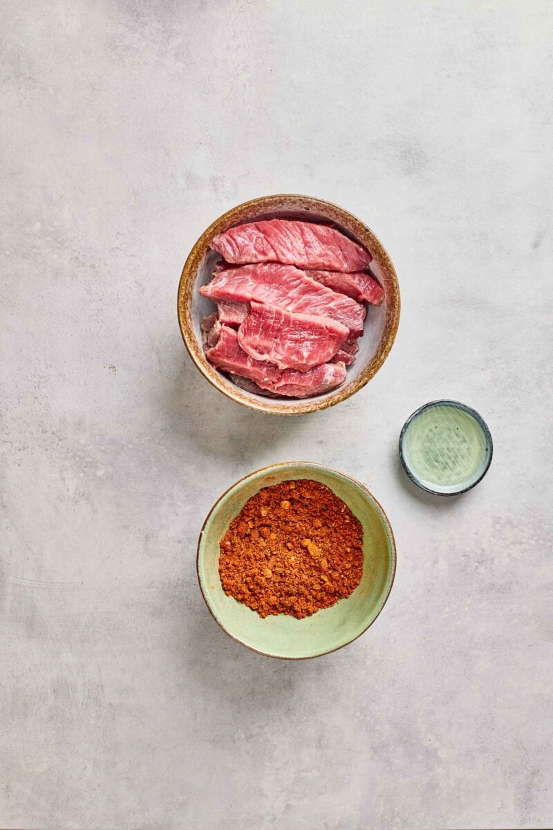 A bowl of raw sliced beef, a bowl of dry spice mix, and a small bowl of clear liquid on a light surface.